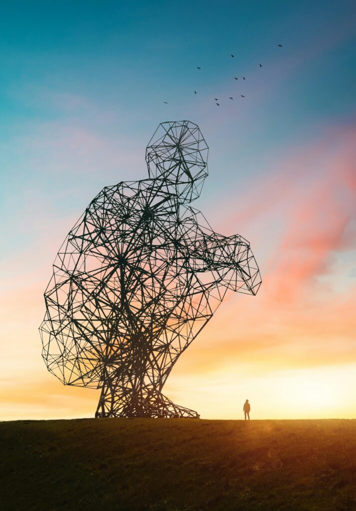 Person standing beside a large modern sculpture of a kneeling person with a sun setting behind them.