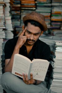 Man in hat sitting reading a book, surrounded by stacks of books